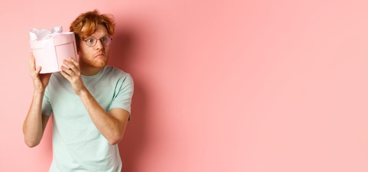 Love and holidays concept. Intrigued redhead guy press ear to box and shaking gift, guessing what inside, standing over pink background.