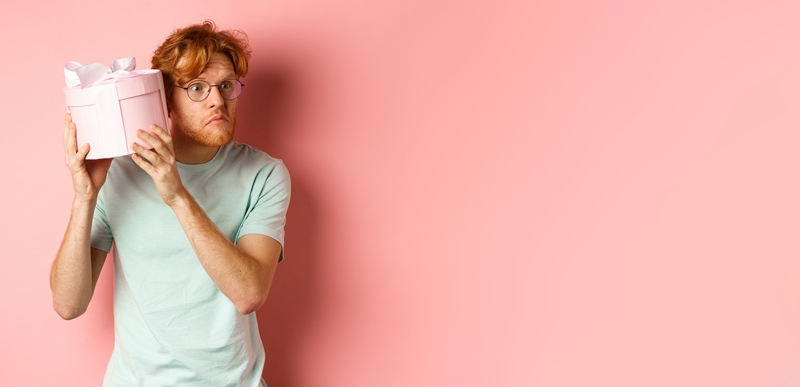 Love and holidays concept. Intrigued redhead guy press ear to box and shaking gift, guessing what inside, standing over pink background.
